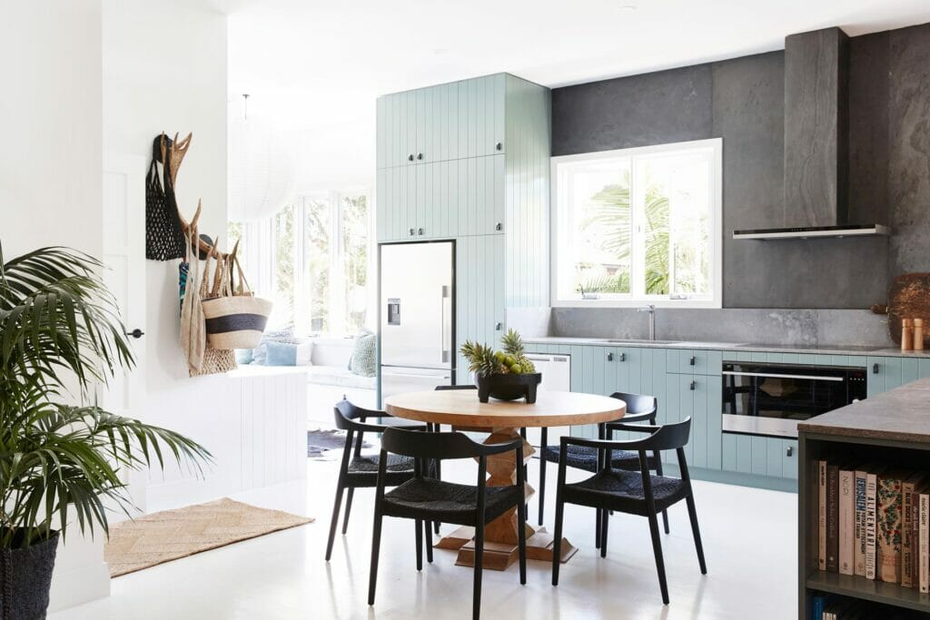 Modern caesarstone kitchen and living area with green cabinetry, grey stone splashback, concrete benchtops and round wooden dining table with black chairs.