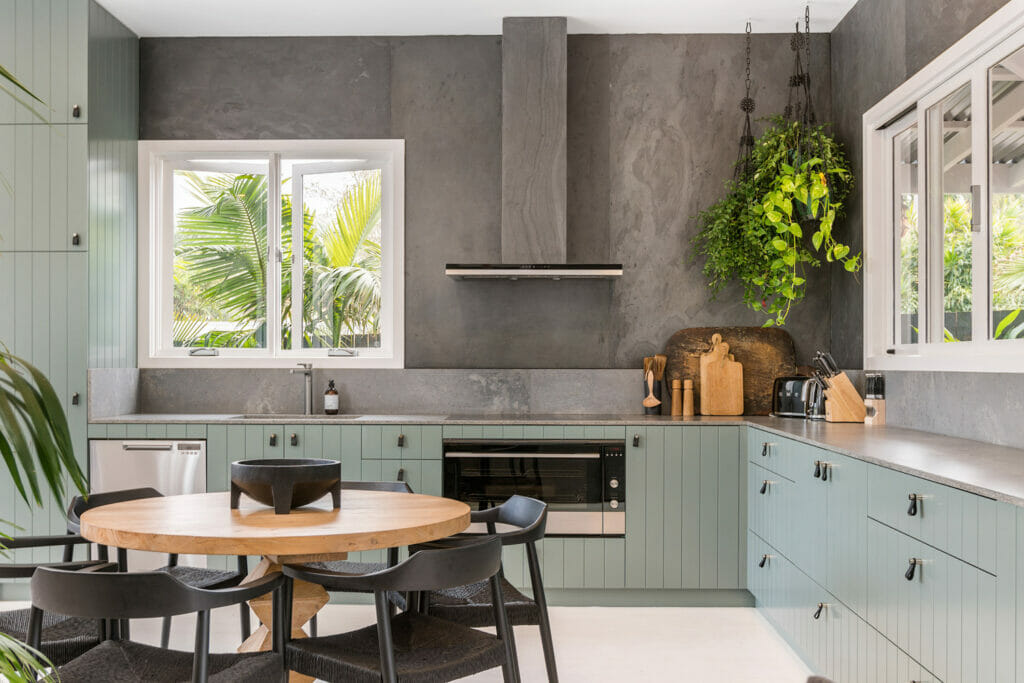 Modern caesarstone kitchen and living area with green cabinetry, grey stone splashback, concrete benchtops and round wooden dining table with black chairs.