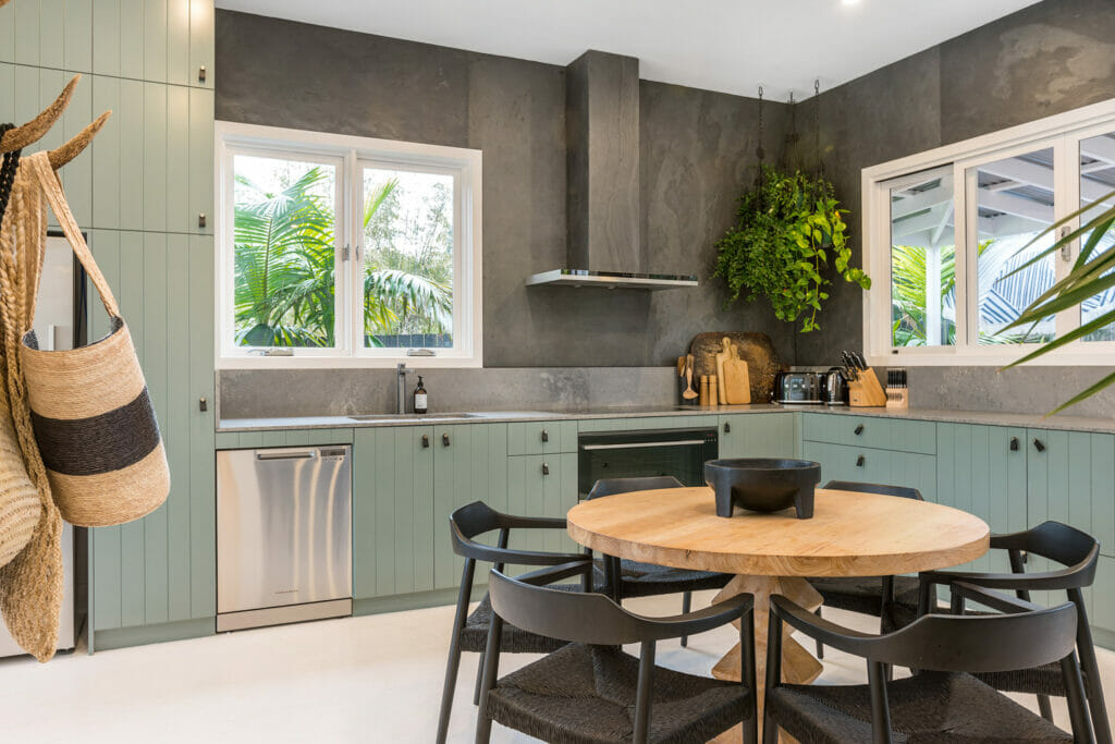 Modern caesarstone kitchen and living area with green cabinetry, grey stone splashback, concrete benchtops and round wooden dining table with black chairs.