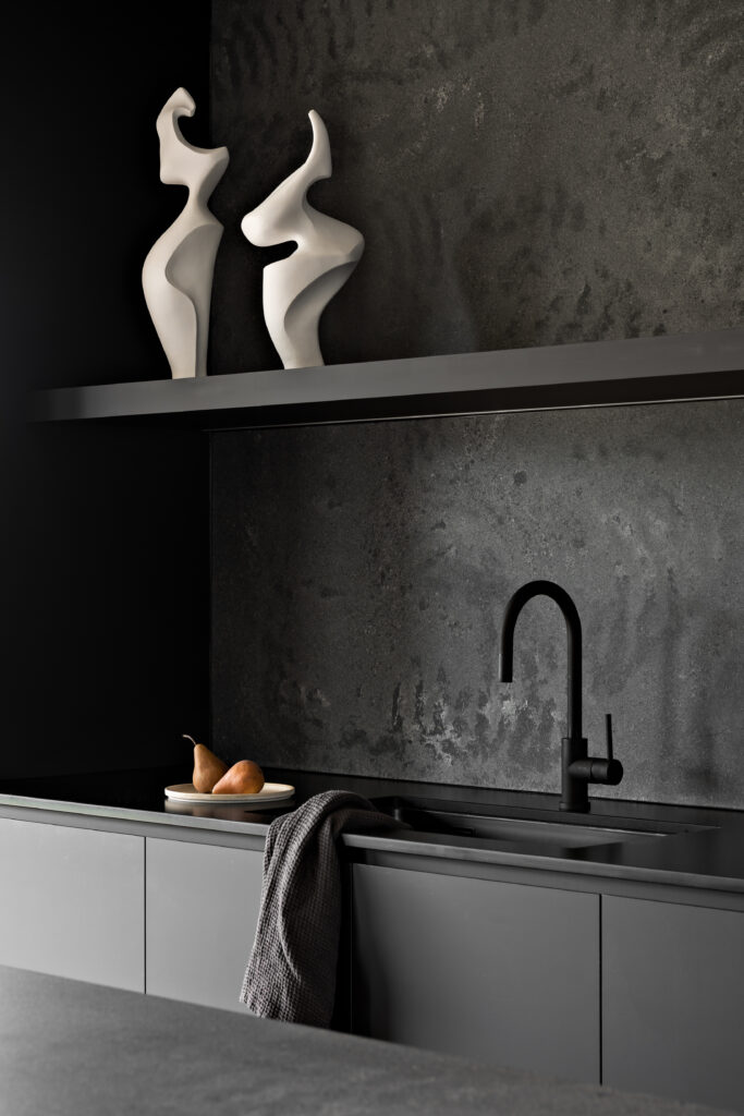 Close-up of matte black kitchen joinery with integrated sink, textured dark splashback, and sculptural white decor on open shelf.