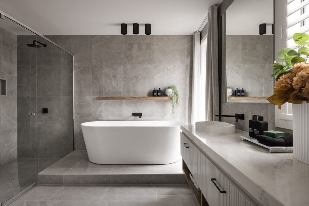 Contemporary bathroom with freestanding bathtub, large grey tiles, timber shelf, and Caesarstone<sup>®</sup> vanity benchtop.