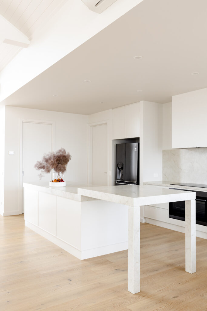 Contemporary kitchen featuring Caesarstone<sup>®</sup> island with custom leg split-level detail, full-height splashback, and integrated white joinery against pale oak flooring.