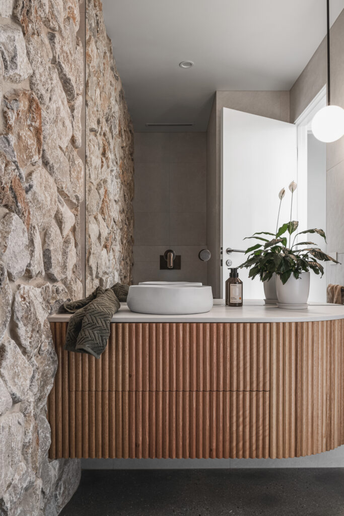 Powder room vanity in Caesarstone<sup>®</sup> with ribbed timber cabinetry, stone feature wall, and round basin.