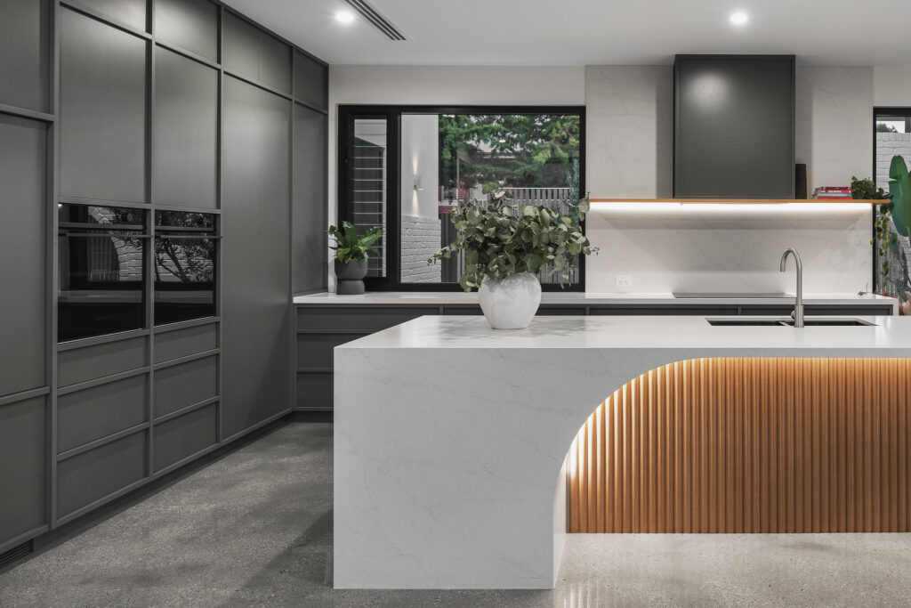 Kitchen island in Caesarstone<sup>®</sup> with curved ribbed timber detail, paired with dark cabinetry and integrated appliances.