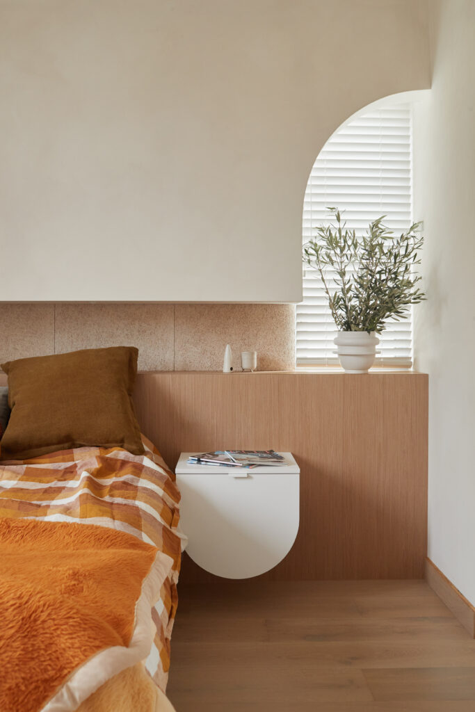 Minimalist bedroom with warm timber accents, curved wall niche and soft natural light, featuring a floating bedside table and layered textiles.