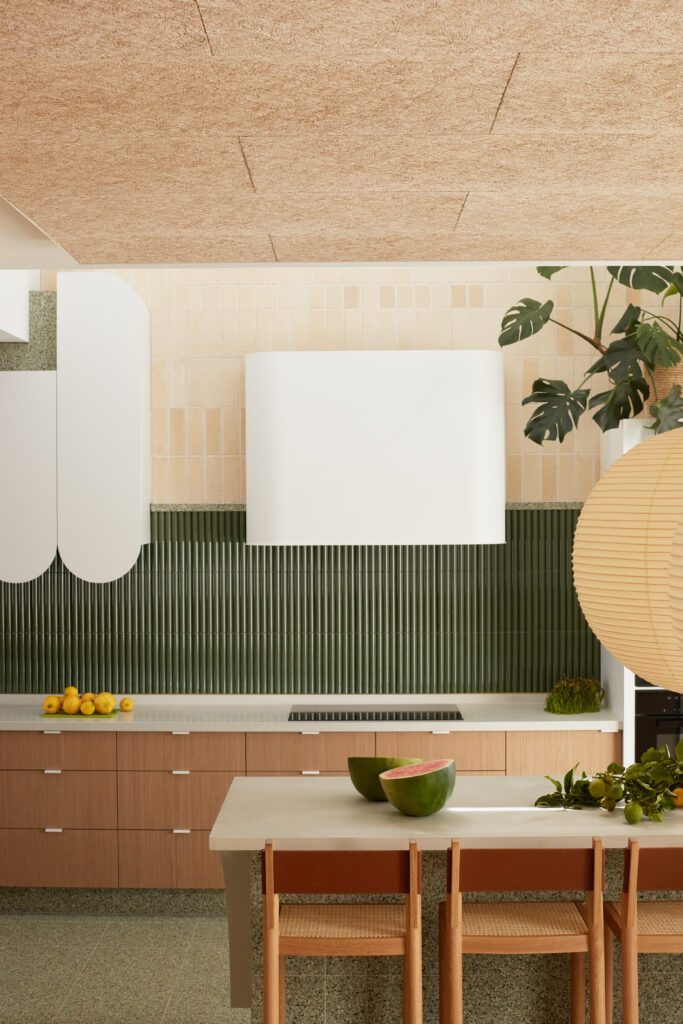 Kitchen view of Caesarstone<sup>®</sup> ICON™ benchtop and island, oak joinery, fluted green splashback, and soft natural light across mid-century inspired finishes.