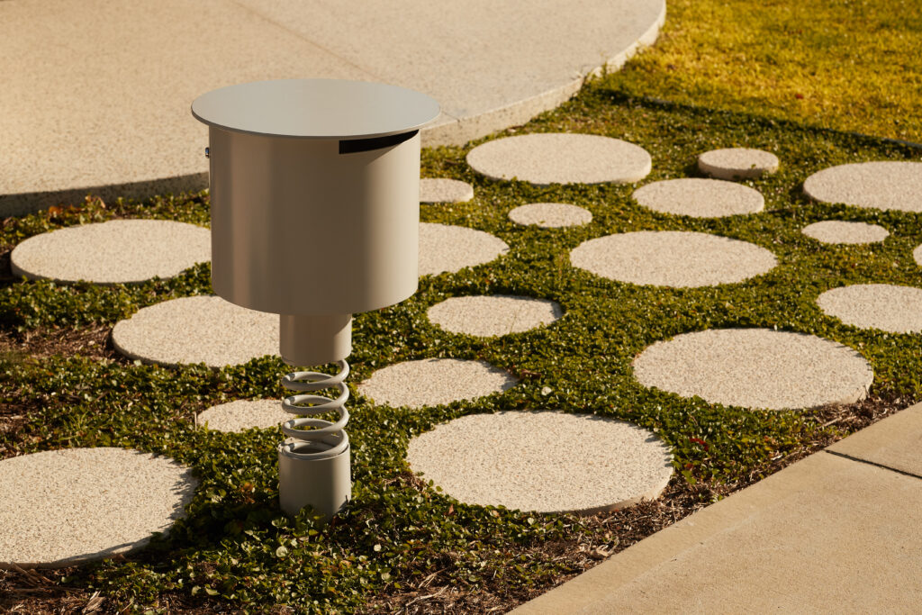 Custom letterbox by Neil Cownie, perched playfully on a suspension spring among circular concrete pavers and creeping groundcover.