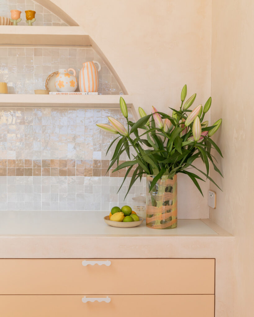 Close-up of a modern Australian kitchen featuring Caesarstone<sup>®</sup> Porcelain White Ciment benchtop, soft peach cabinetry, tiled splashback, and styled open shelving with decorative ceramics and fresh flowers.