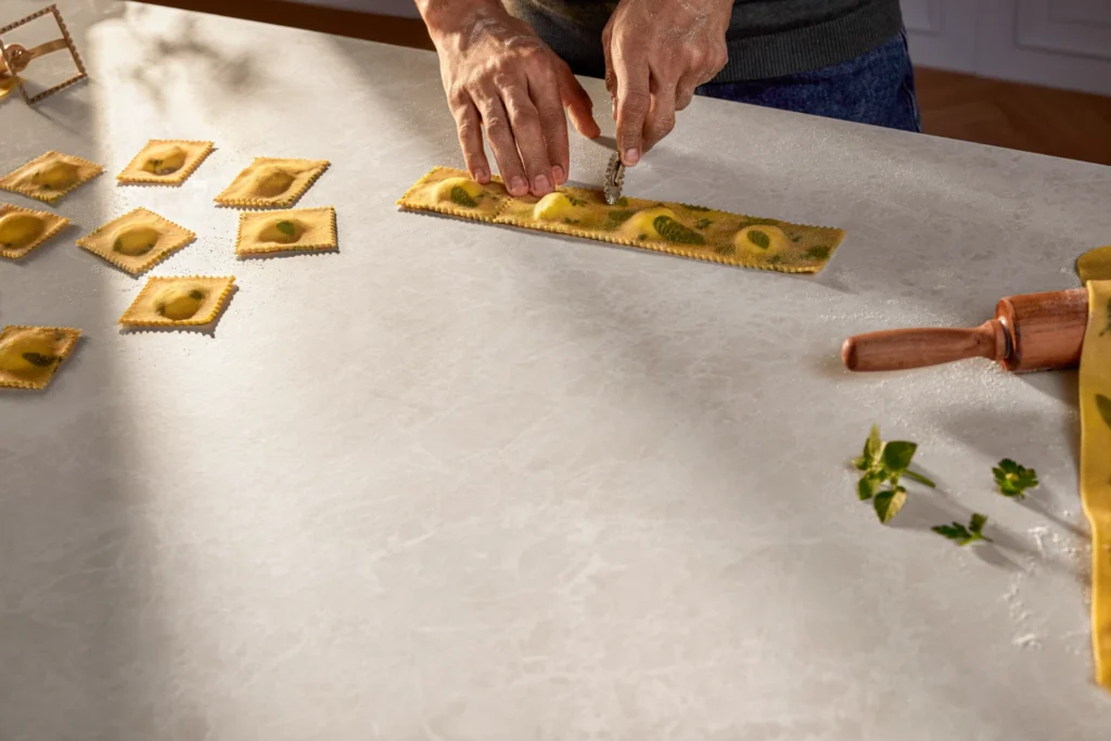 Hands preparing fresh pasta on a Caesarstone<sup>®</sup> porcelain benchtop, showcasing a soft neutral surface in a kitchen setting