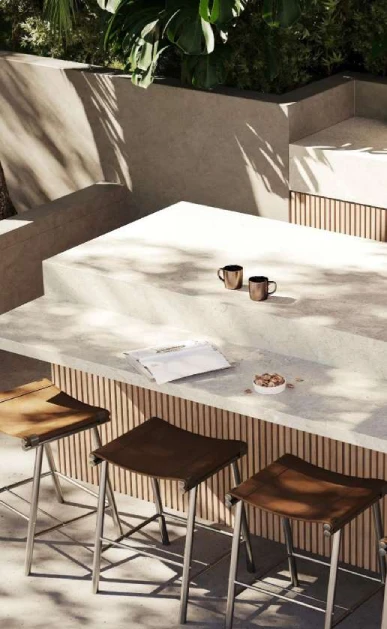 Caesarstone<sup>®</sup> Fossillia™ 545 porcelain benchtop in alfresco entertaining zone with slatted timber joinery, bar stools and garden backdrop