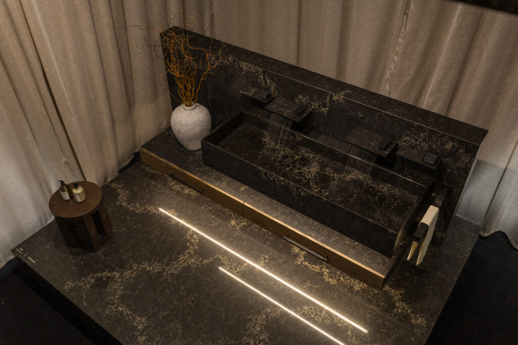 Freestanding bath clad in Caesarstone<sup>®</sup> ICON™ Vanilla Noir, illuminated from below and framed by layered textiles, evoking a calming, immersive environment.