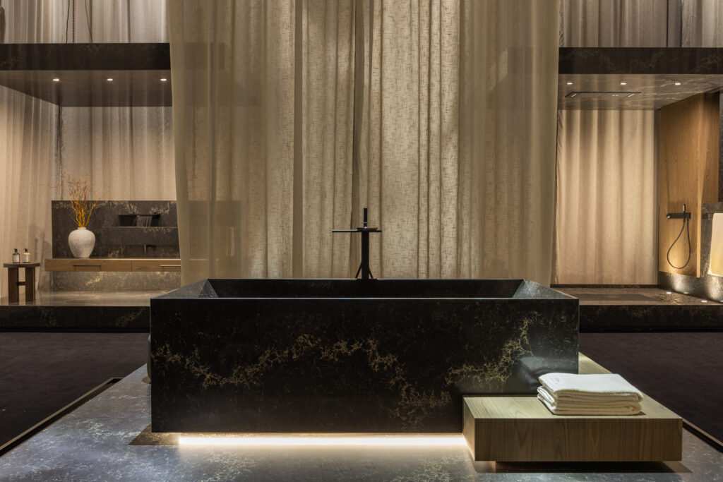 Freestanding bath clad in Caesarstone<sup>®</sup> ICON™ Vanilla Noir, illuminated from below and framed by layered textiles, evoking a calming, immersive environment.