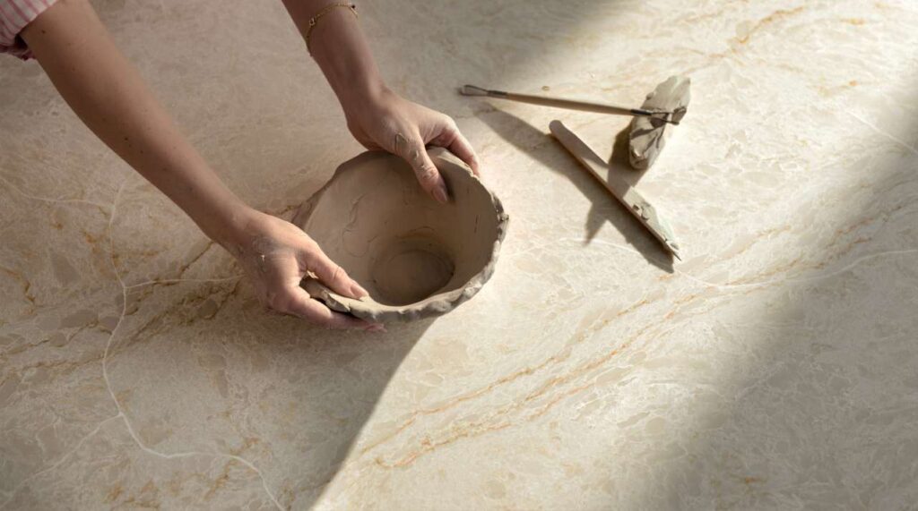 Caesarstone<sup>®</sup> Taj Whisper™ 8251 ICON Mineral benchtop surface detail with hands shaping clay bowl, tools and soft natural light highlighting feather-light veining