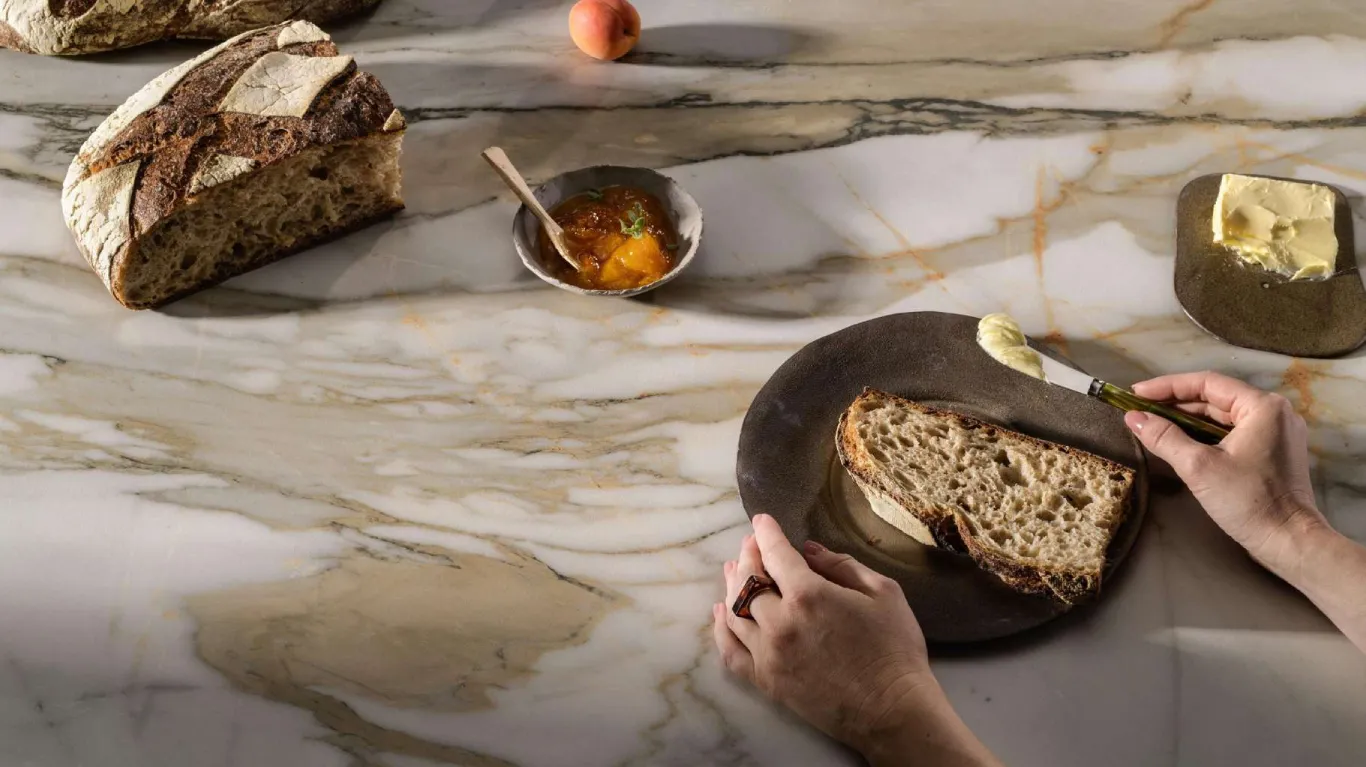 Caesarstone<sup>®</sup> Antikella™ 536 porcelain benchtop with gold veining, styled with sourdough bread and butter in a contemporary kitchen setting