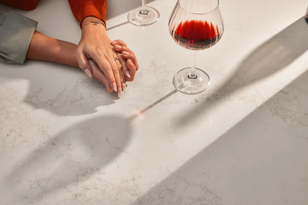 A hand resting on a Caesarstone<sup>®</sup> ICON™ Solenna benchtop alongside two wine glasses, one filled with red wine