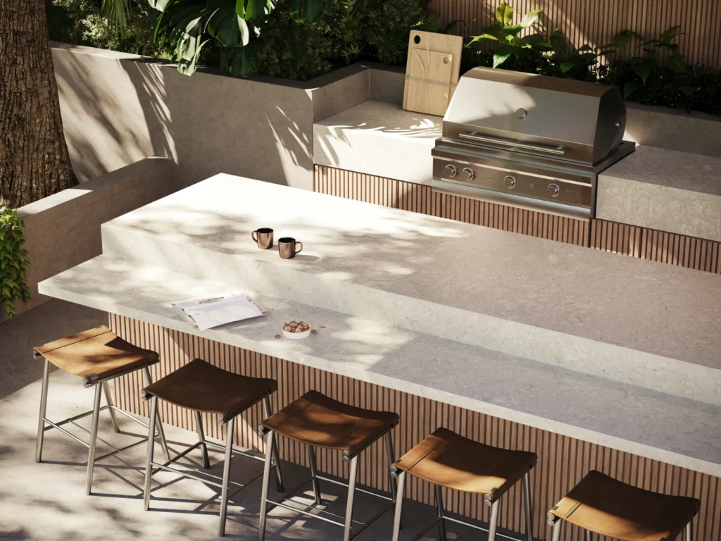 Pale warm stone-looking Caesarstone<sup>®</sup> Porcelain benchtop in a contemporary outdoor alfresco kitchen, with timber bar stools, an integrated grill and battened timber detailing.