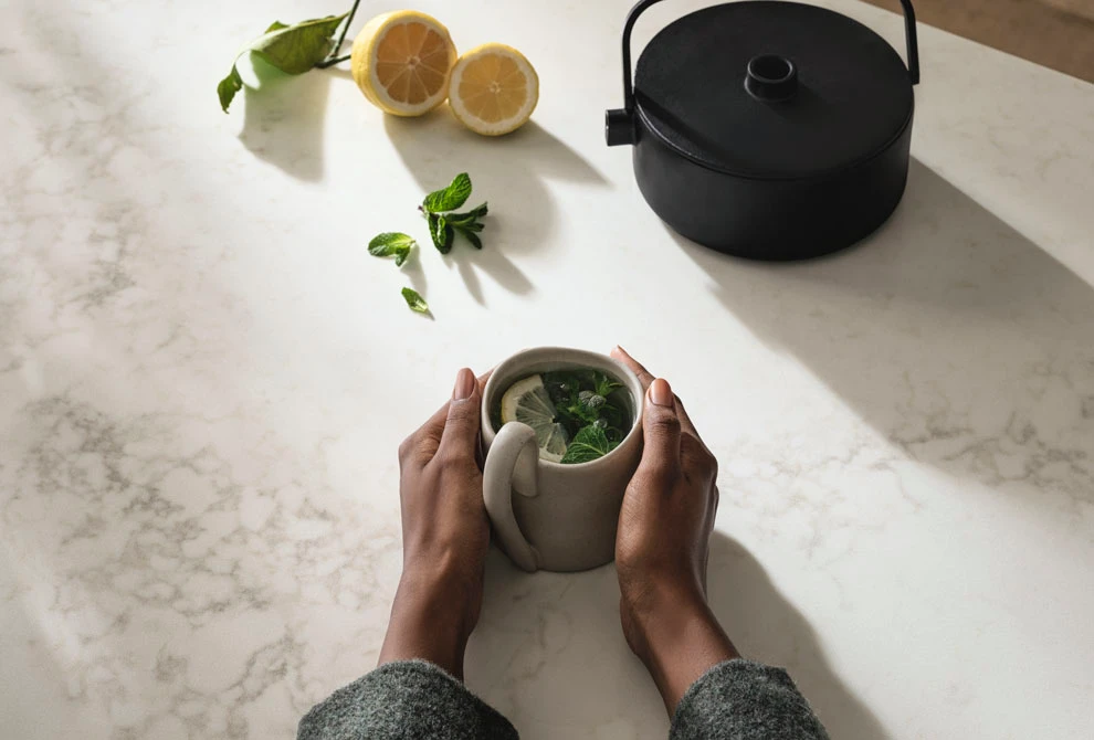 Hands holding a stone matcha bowl on a Caesarstone<sup>®</sup> ICON™ Calacatta Lacebound benchtop, styled with lemons and a cast iron teapot