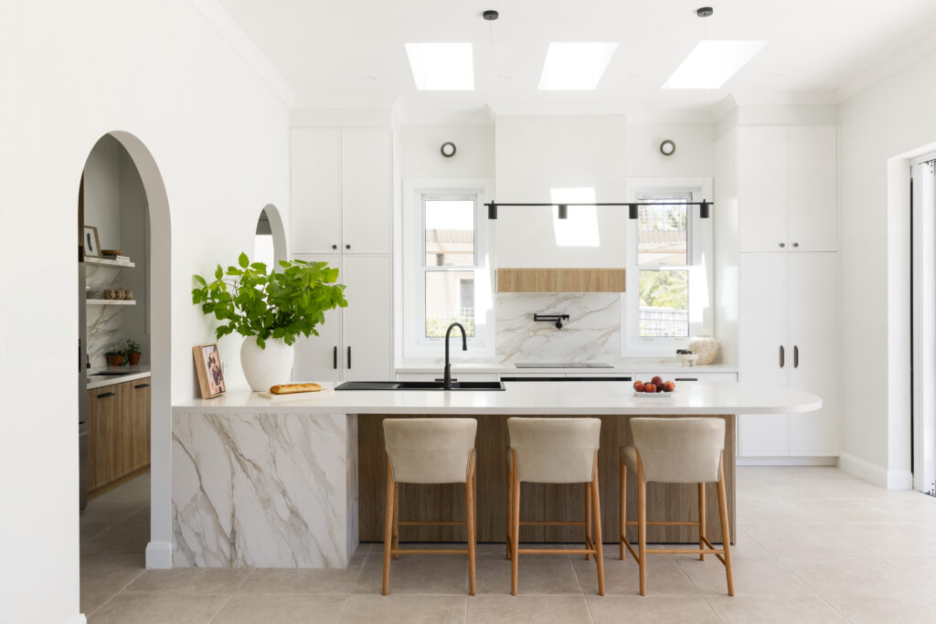 Open-plan kitchen featuring Caesarstone<sup>®</sup> Mirabel™ porcelain and ICON Organic white benches, waterfall island and splashback, timber joinery, arched pantry entry and skylit ceiling in Seaforth home by Heliconia