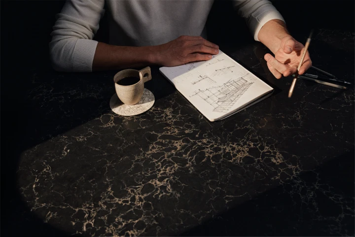 Dark marble-look Caesarstone<sup>®</sup> benchtop with gold veining in a moody, low-lit room, styled with a sketchbook, pencil and espresso cup during kitchen planning.