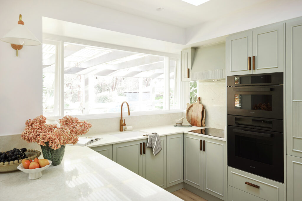 Coastal kitchen featuring sage green cabinetry, copper fittings, integrated black appliances and a Caesarstone<sup>®</sup> Sedara benchtop.