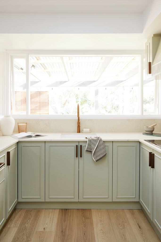 U-shaped coastal kitchen with sage cabinetry, copper tapware, a white sink and a Caesarstone<sup>®</sup> Sedara benchtop beneath a picture window.