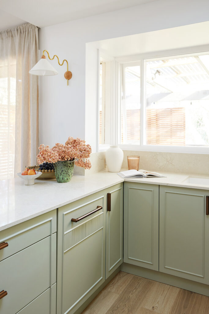 Sage green kitchen corner with fluted joinery, copper handles, a brass wall light and a Caesarstone<sup>®</sup> Sedara benchtop.
