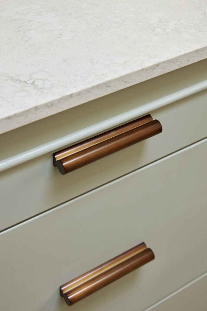 Warm copper cabinet handles on sage green drawers beneath a Caesarstone<sup>®</sup> Sedara benchtop.