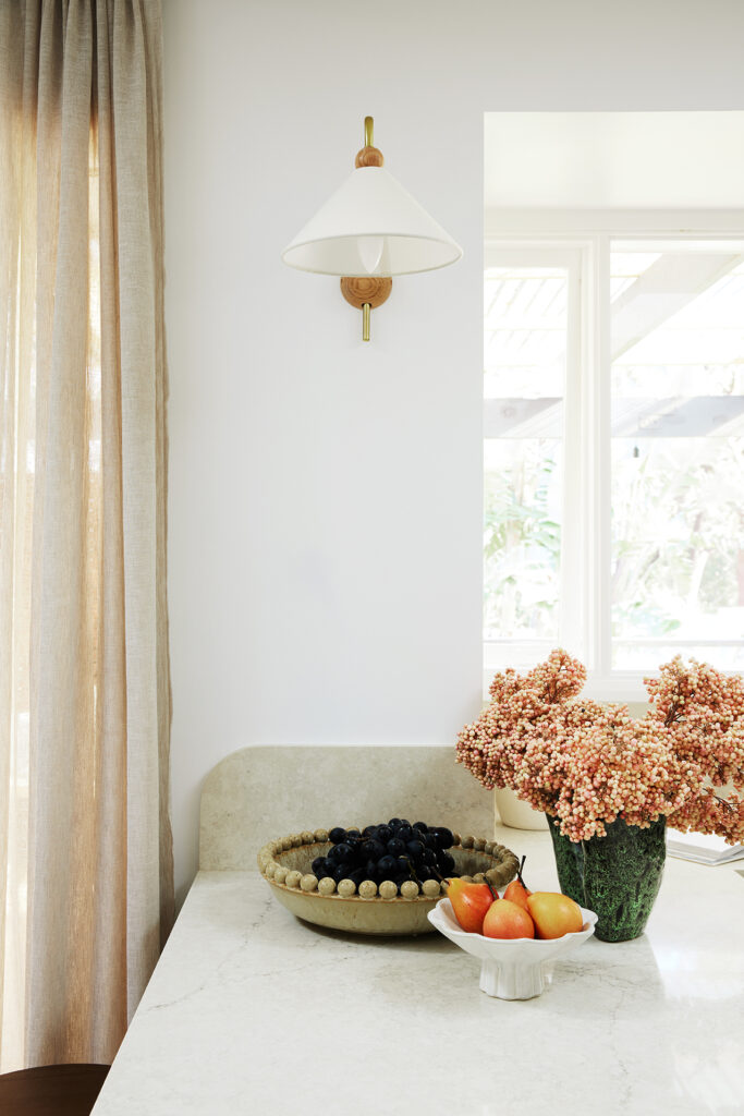 Caesarstone<sup>®</sup> Sedara benchtop styled with a bowl of grapes, pears and dried botanicals beneath a wall sconce and soft linen curtains.
