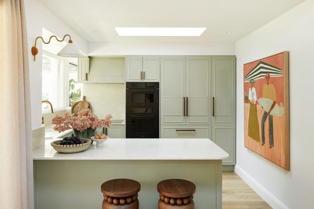 Renovated coastal kitchen with sage green cabinetry, a Caesarstone<sup>®</sup> Sedara island benchtop, timber bar stools and a coastal artwork.