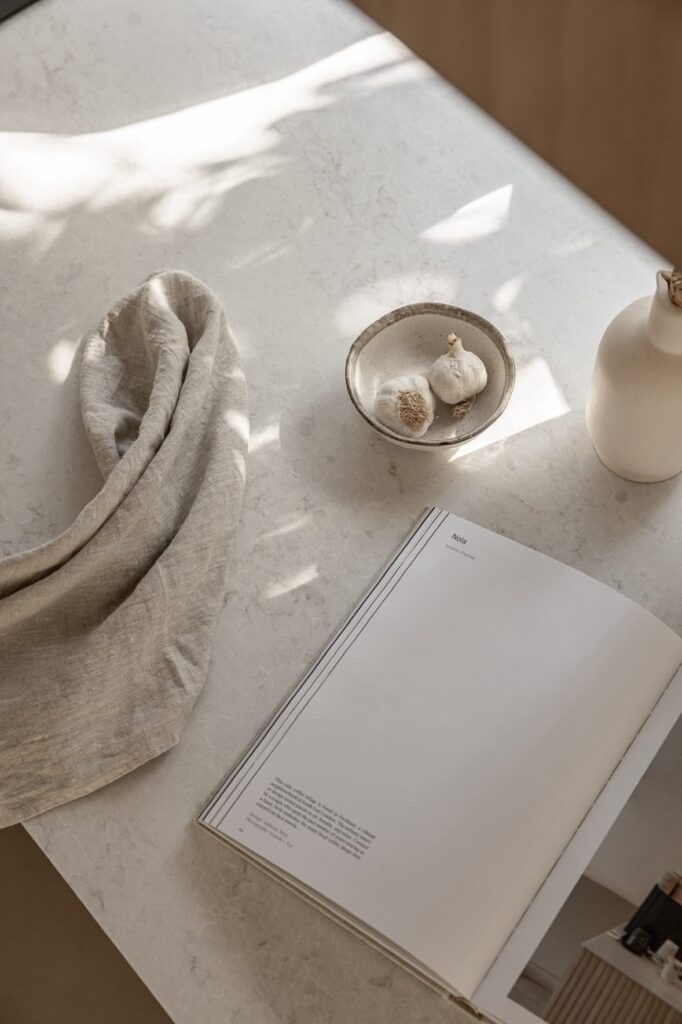 Styled detail of Caesarstone Lunar Frost benchtop with linen cloth, bowl of garlic and open book.