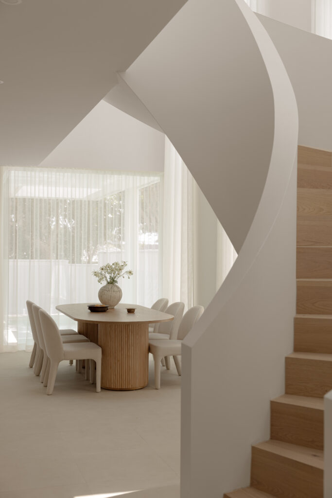Round timber dining table beneath a curved staircase, framed by sheer curtains.