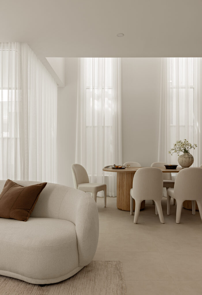 Curved bouclé sofa in a living room with sheer curtains and timber dining table beyond.