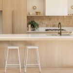 Caesarstone 2141 Snow surface in Minimalist kitchen with wooden cabinetry, white island, two stools, modern faucet, and natural light.