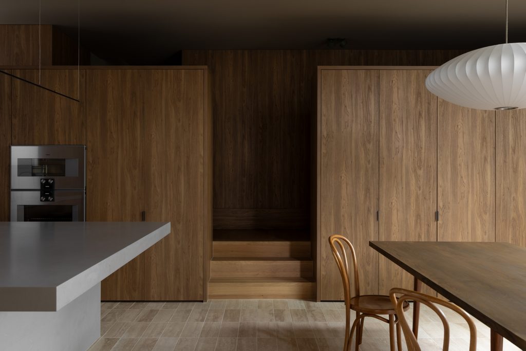 Kitchen and dining area with oak-toned timber panelling, a Caesarstone<sup>®</sup> Raw Concrete benchtop and a round pendant light above a timber dining table.