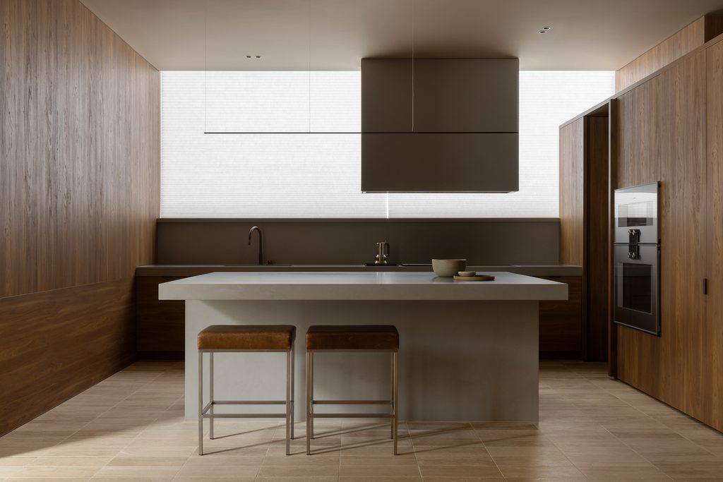 Symmetrical kitchen island featuring a Caesarstone<sup>®</sup> Raw Concrete benchtop, flanked by leather bar stools, timber joinery and a rectangular pendant light.