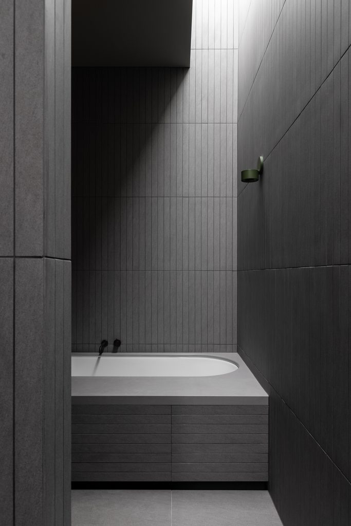 Dark, ribbed-tile bathroom with a sunken bath set into a Caesarstone<sup>®</sup> Raw Concrete surround, lit from a skylight above.