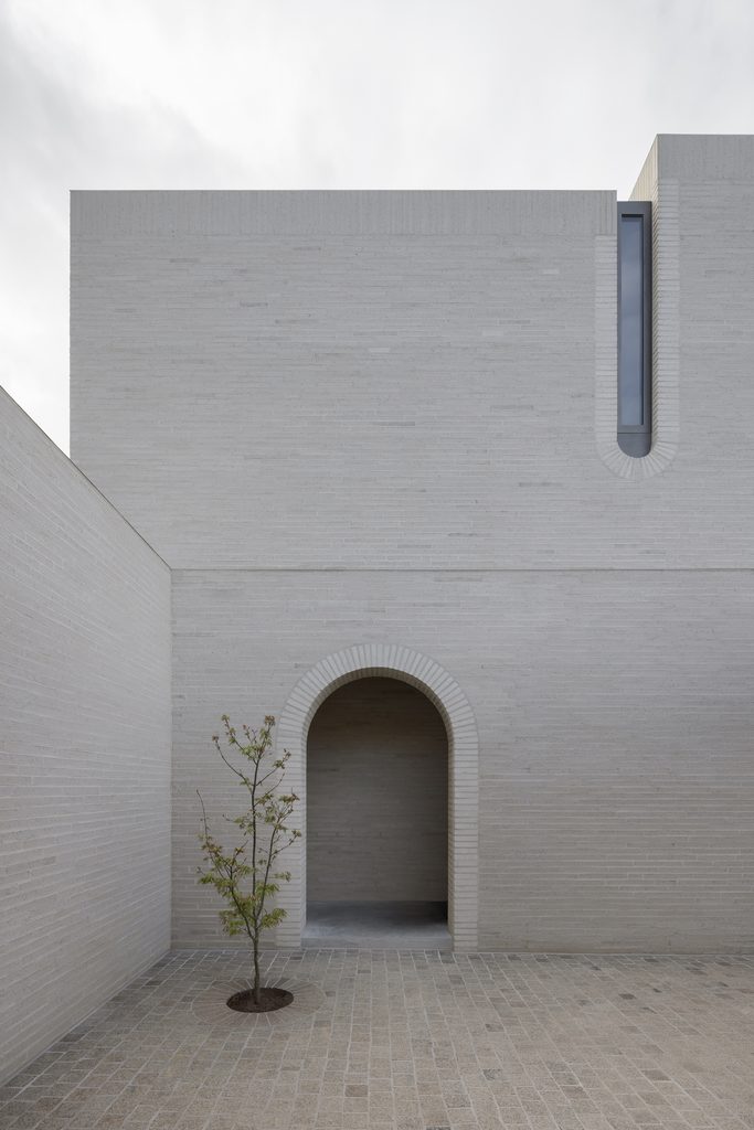 Pale brick courtyard at Casa Piva with an arched doorway and a single young tree, seen against an overcast sky.